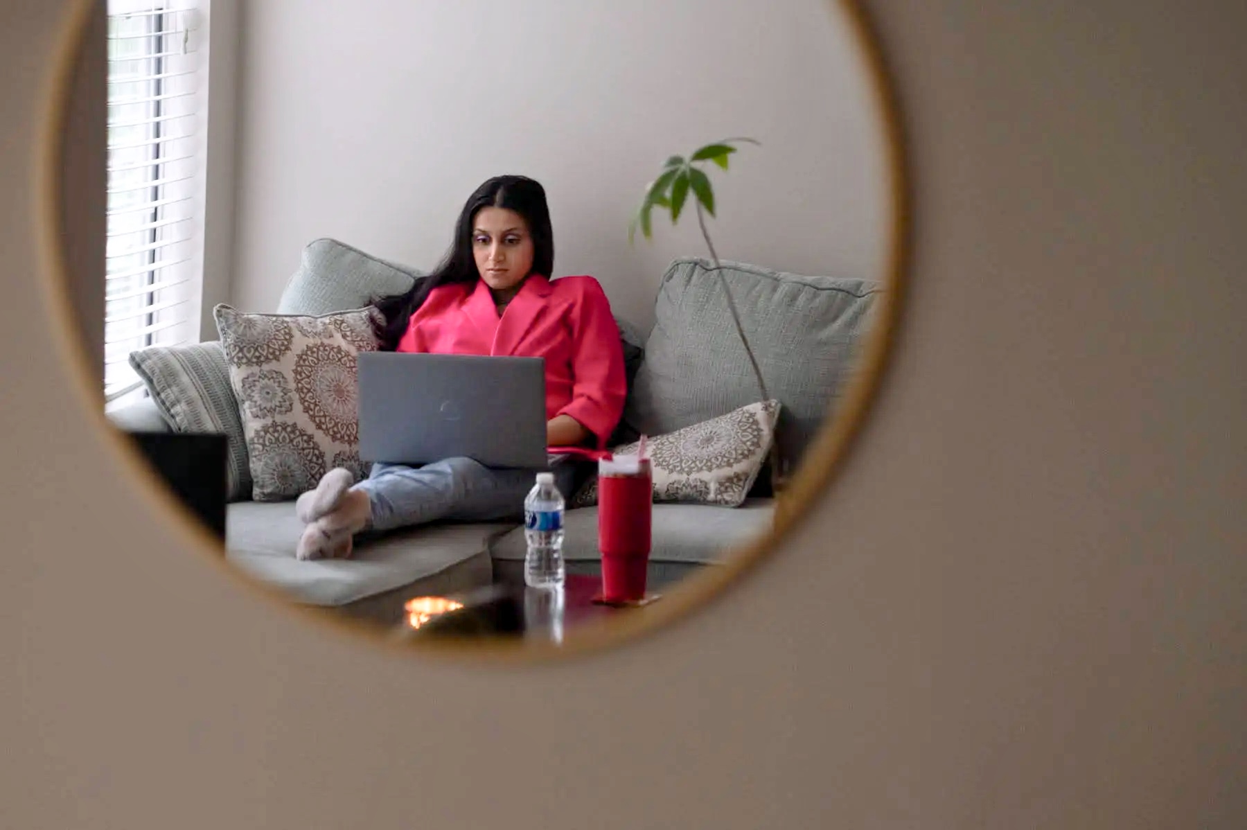 Seen in the reflection of a round wall mirror, a woman with long dark hair sits on a couch, a laptop open on her lap and her legs stretched out in front of her. A water bottle and a tall red tumbler stand on the table before her.
