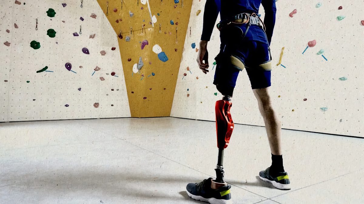 Climber with a prosthetic leg at an indoor climbing wall A climber with a red prosthetic leg stands in a harness at an indoor climbing wall, seen from behind and from the shoulders down, facing colored holds and a yellow wall section.