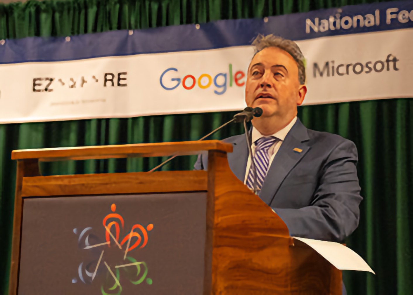 An adult Man who is Blind speaks into a microphone at a podium, holding papers, with convention banners and a green curtain behind him.