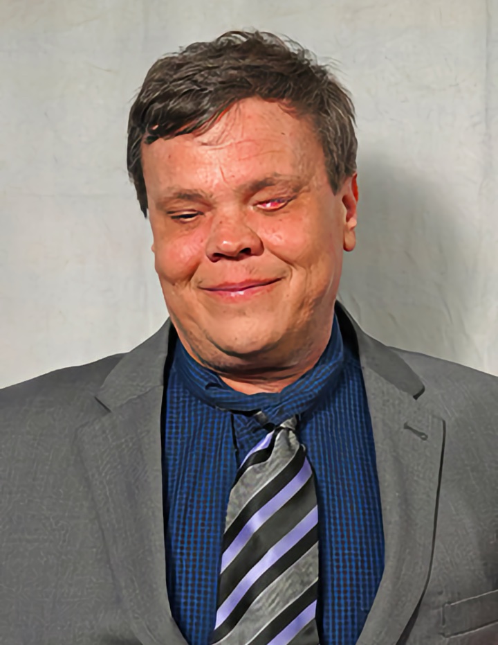 A man with short dark hair is smiling gently, wearing a gray suit jacket, a dark blue checkered shirt, and a black, gray, and lavender striped tie against a light neutral backdrop.