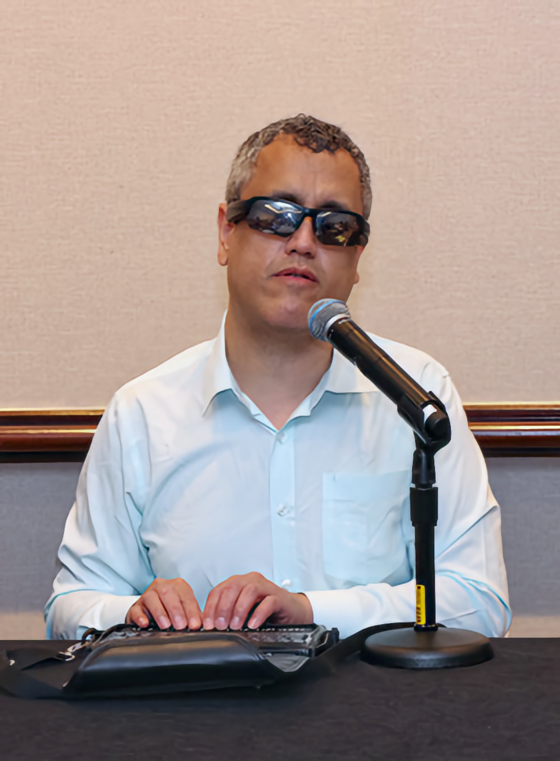 A middle aged man sits at a dark conference table wearing dark tinted glasses. His hands rest on a refreshable Braille display in front of a tabletop microphone.