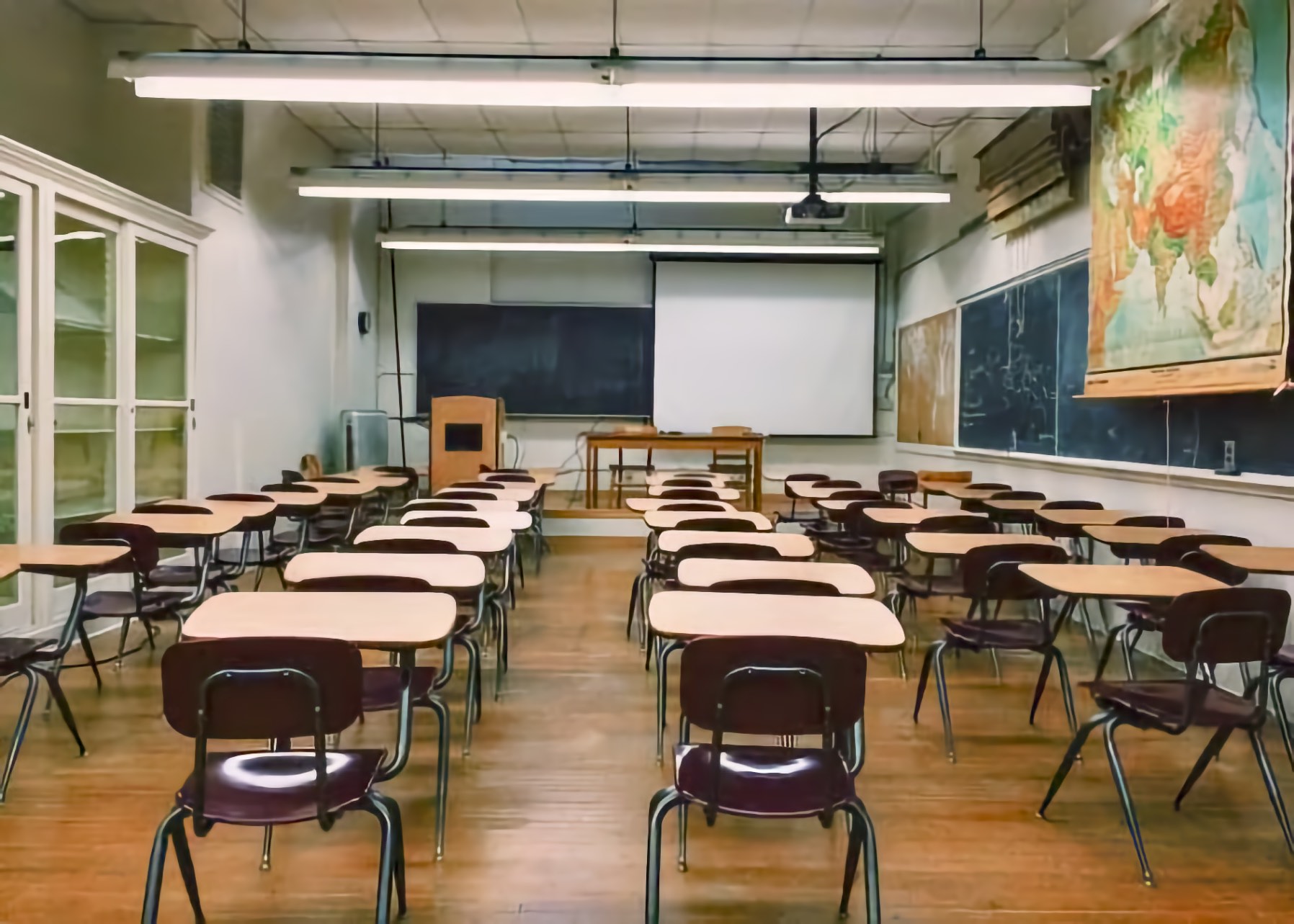 Empty classroom with rows of wooden student desks and dark chairs facing a chalkboard and a projection screen, with a world map on the right wall.