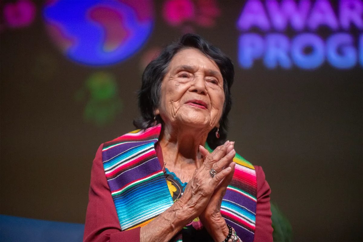 A woman smiles softly with her palms pressed together at her chest, in front of a glowing projected backdrop.