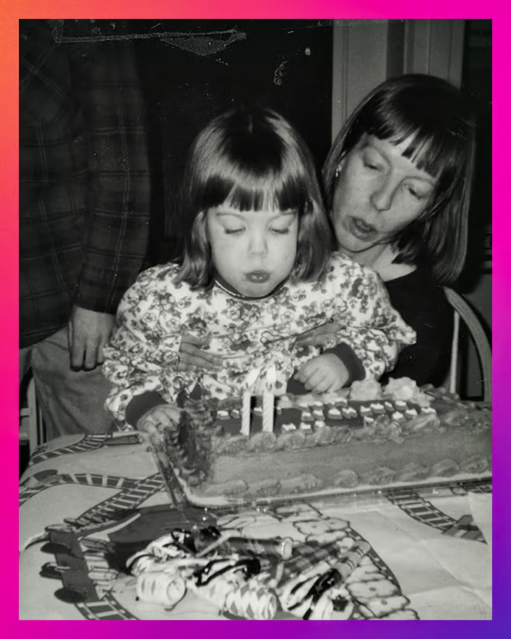 A child in a floral dress leans forward to blow out two candles on a rectangular birthday cake while a woman behind the child leans forward as if assisting. The table is decorated with a train-themed tablecloth and party items.