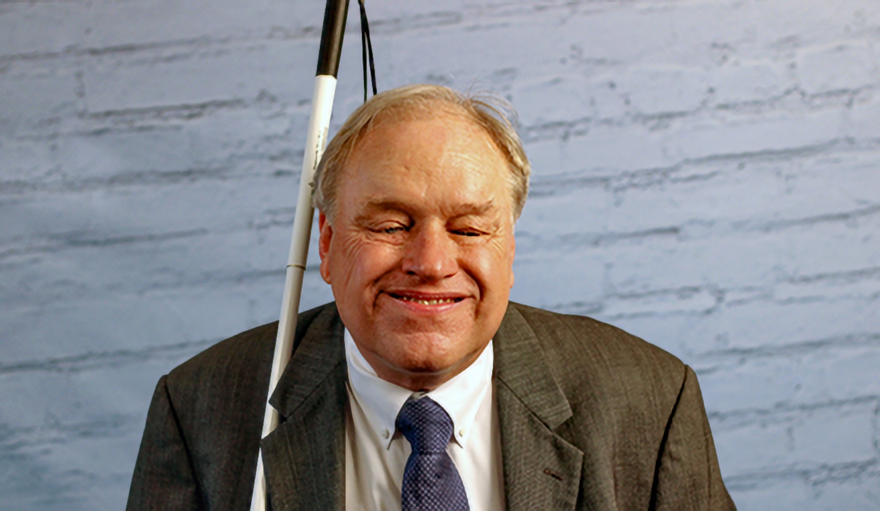 A Blind man smiling broadly with his eyes closed, a long white cane rising past his shoulder in front of a white painted brick wall.