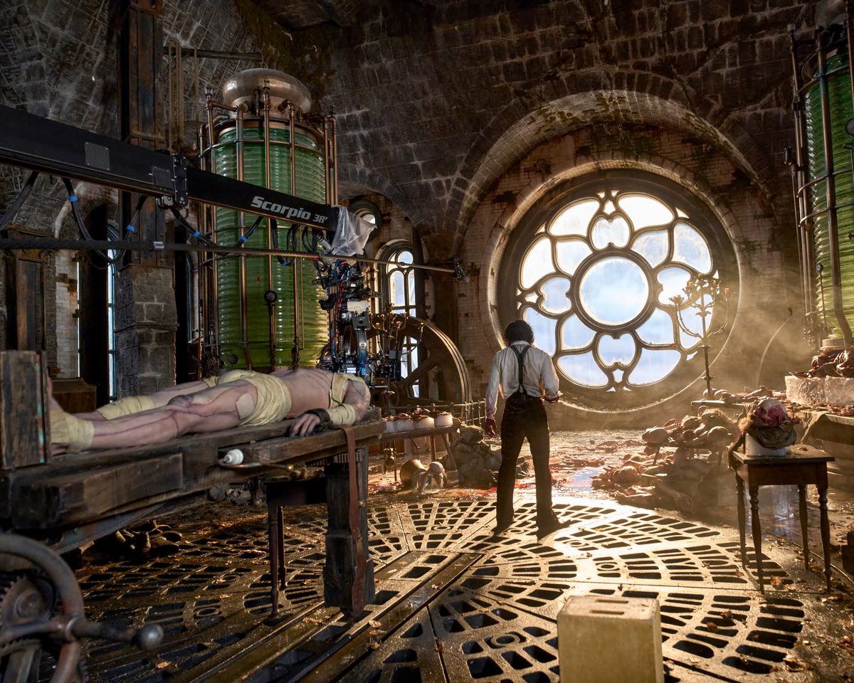 Stone laboratory with a large round window and green glass tanks. A person seen from behind stands near a bandaged figure lying on a table, under a film camera on a crane arm.