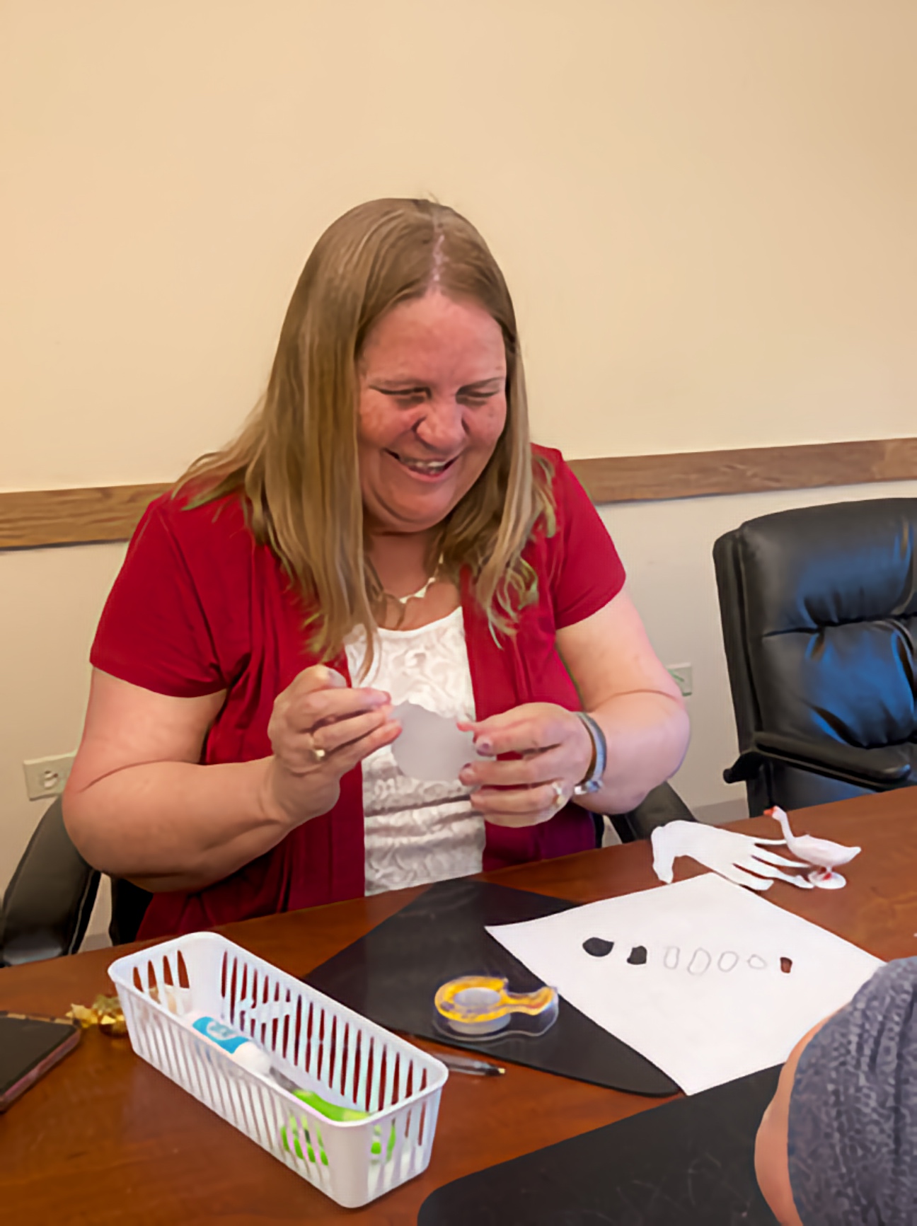 Julie Deden smiles as she cuts a paper shape at a table with art supplies.