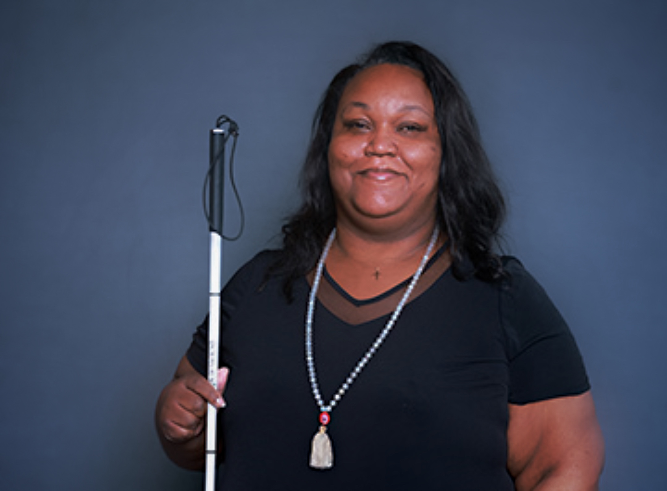 A smiling Black woman with long, wavy hair holds a white cane against a dark blue-gray background. She wears a black top and a beaded necklace.