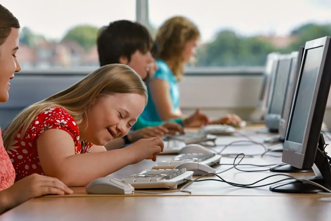 A girl with a disability laughs as she types at a keyboard in a row of computers by a window, with three other children working at screens along the same desk.